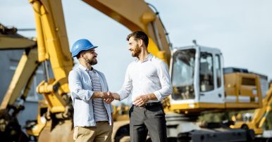 Man In A Hard Hat Shaking Hands With A Business Owner With Heavy Equipment In The Background